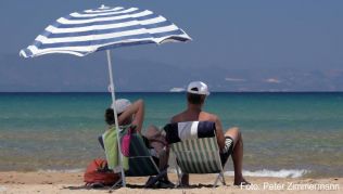 Touristen sitzen am Strand bei Loutra Kilini im Westen der Halbinsel Peloponnes in Griechenland unter einem weißblauen Sonnenschirm##Foto: Peter Zimmermann Touristen sitzen am Strand bei Loutra Kilini im Westen der Halbinsel Peloponnes in Griechenland unter einem weißblauen Sonnenschirm
