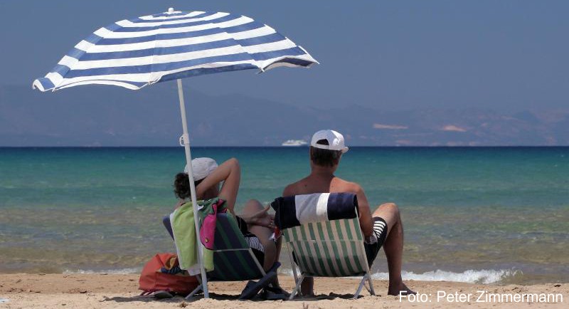 Touristen sitzen am Strand bei Loutra Kilini im Westen der Halbinsel Peloponnes in Griechenland unter einem weißblauen Sonnenschirm