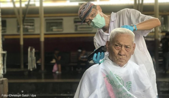 Auf dem Hauptbahnhof Hua Lamphong schneidet ein Friseur einem älteren Mann die Haare. Der Haarschnitt ist kostenlos